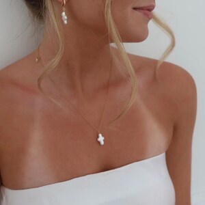 May include: A woman wearing a white strapless top with a delicate gold chain necklace featuring a white pearl cross pendant.