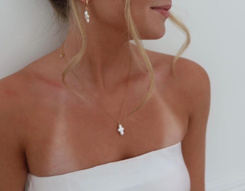 May include: A woman wearing a white strapless top with a delicate gold chain necklace featuring a white pearl cross pendant.