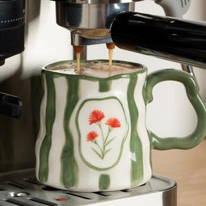 May include: A stainless steel ASD espresso machine with a black bean hopper and portafilter, dispensing coffee into a white ceramic mug featuring green vertical stripes and a central illustration of three small orange flowers.