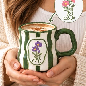 May include: A white ceramic mug with a wavy, textured surface and vertical dark green stripes. The front features a central oval panel containing a purple flower illustration and the letter A. An inset shows a pink flower illustration with the letter B.