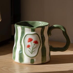May include: A ceramic mug with a wavy, irregular rim and handle, featuring vertical green stripes on a white background. A central oval frame on the front displays a hand-painted illustration of three small red flowers with green stems.