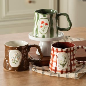 May include: Three ceramic mugs with irregular, wavy rims and handles. One mug is green with white stripes and red flowers, one is brown with white dots and white daisies, and one is white with a red plaid pattern and green lily of the valley.
