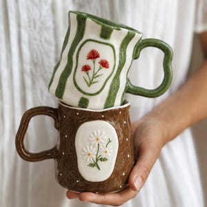 May include: Two ceramic mugs with wavy handles. The top mug is white with vertical green stripes and a central oval featuring red flowers. The bottom mug is brown with white polka dots and a central oval featuring white daisy-like flowers.
