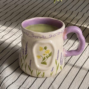 May include: A ceramic mug with a wavy lavender handle and rim, featuring a cream-colored body decorated with painted purple lavender sprigs and a central embossed panel displaying a cluster of yellow flowers, filled with a light green liquid.