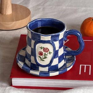 May include: A ceramic mug and matching saucer set featuring a blue and white checkered pattern. The mug has a wavy rim and a curved handle, with a central oval emblem displaying two red roses with green leaves. The mug is filled with dark liquid.