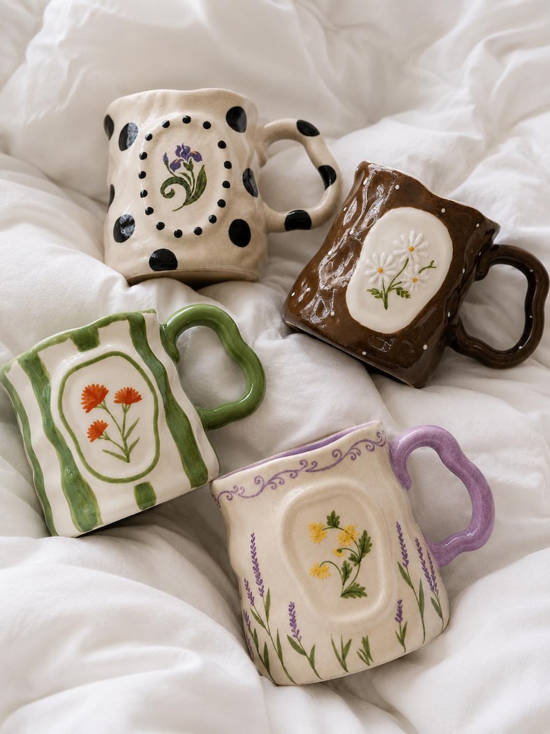 May include: Four ceramic mugs with irregular, wavy shapes and handles. One is cream with black polka dots and a purple flower, one is dark brown with white daisies, one is white with green stripes and orange flowers, and one is cream with lavender.