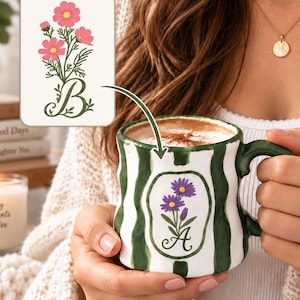 May include: A white ceramic mug with vertical dark green stripes and a scalloped rim. The front features a central oval emblem containing a purple flower illustration and the letter A. An inset shows a pink flower design with the letter B.