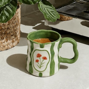 May include: A ceramic mug with a wavy, irregular shape featuring vertical green stripes on a white background. The front displays a central white oval containing a small illustration of three orange flowers. The interior and handle are green.