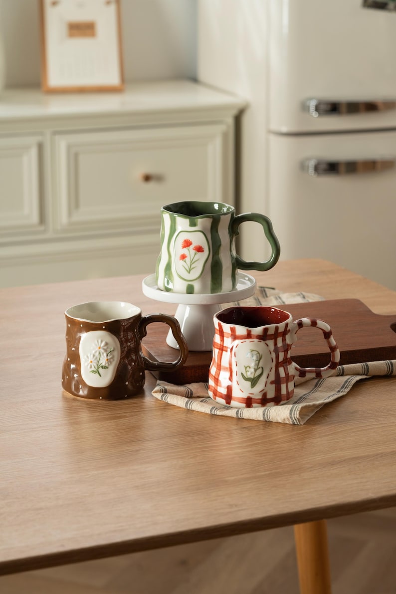 May include: Three ceramic mugs with irregular, wavy rims and handles. One mug is green with white stripes and red flowers, one is brown with white dots and white daisies, and one is white with a red plaid pattern and green lily of the valley.