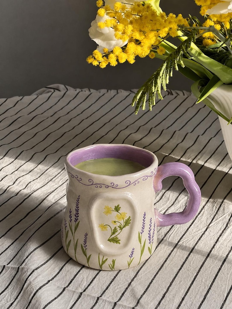 May include: A ceramic mug with a wavy lavender handle and rim, featuring a cream-colored body decorated with painted purple lavender sprigs and a central embossed panel displaying a cluster of yellow flowers, filled with a light green liquid.