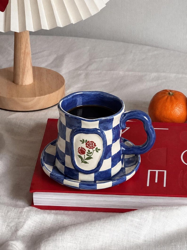May include: A ceramic mug and matching saucer set featuring a blue and white checkered pattern. The mug has a wavy rim and a curved handle, with a central oval emblem displaying two red roses with green leaves. The mug is filled with dark liquid.