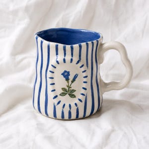 May include: A ceramic mug with a wavy, irregular rim and a curved handle. The exterior features vertical blue stripes on a cream background, with a central oval medallion containing a painted blue gentian flower surrounded by small blue dashes.
