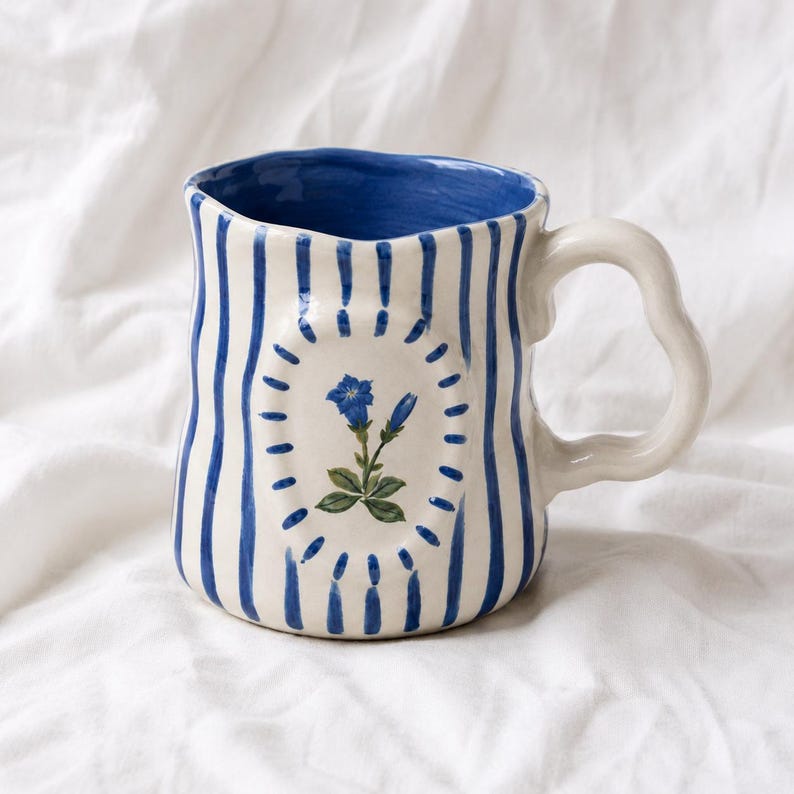 May include: A ceramic mug with a wavy, irregular rim and a curved handle. The exterior features vertical blue stripes on a cream background, with a central oval medallion containing a painted blue gentian flower surrounded by small blue dashes.