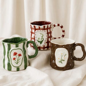 May include: Three ceramic mugs with wavy rims and handles. The left mug is white with green stripes and red flowers. The back mug is white with a red plaid pattern and lily of the valley. The right mug is brown with white dots and daisies.