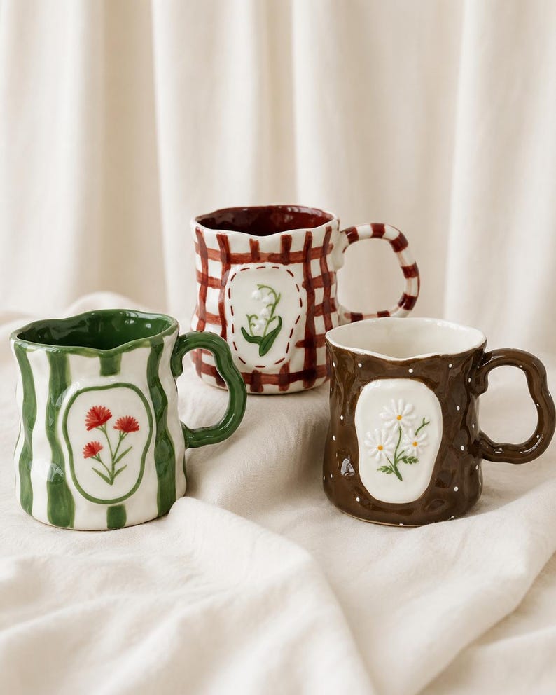May include: Three ceramic mugs with wavy rims and handles. The left mug is white with green stripes and red flowers. The back mug is white with a red plaid pattern and lily of the valley. The right mug is brown with white dots and daisies.