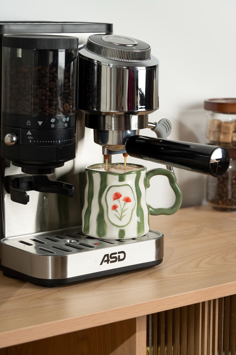 May include: A stainless steel ASD espresso machine with a black bean hopper and portafilter, dispensing coffee into a white ceramic mug featuring green vertical stripes and a central illustration of three small orange flowers.