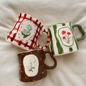 May include: Three ceramic mugs with irregular, wavy rims and handles. One mug features a red gingham pattern with lily of the valley, one has green vertical stripes with red flowers, and one is brown with white polka dots and white daisies.