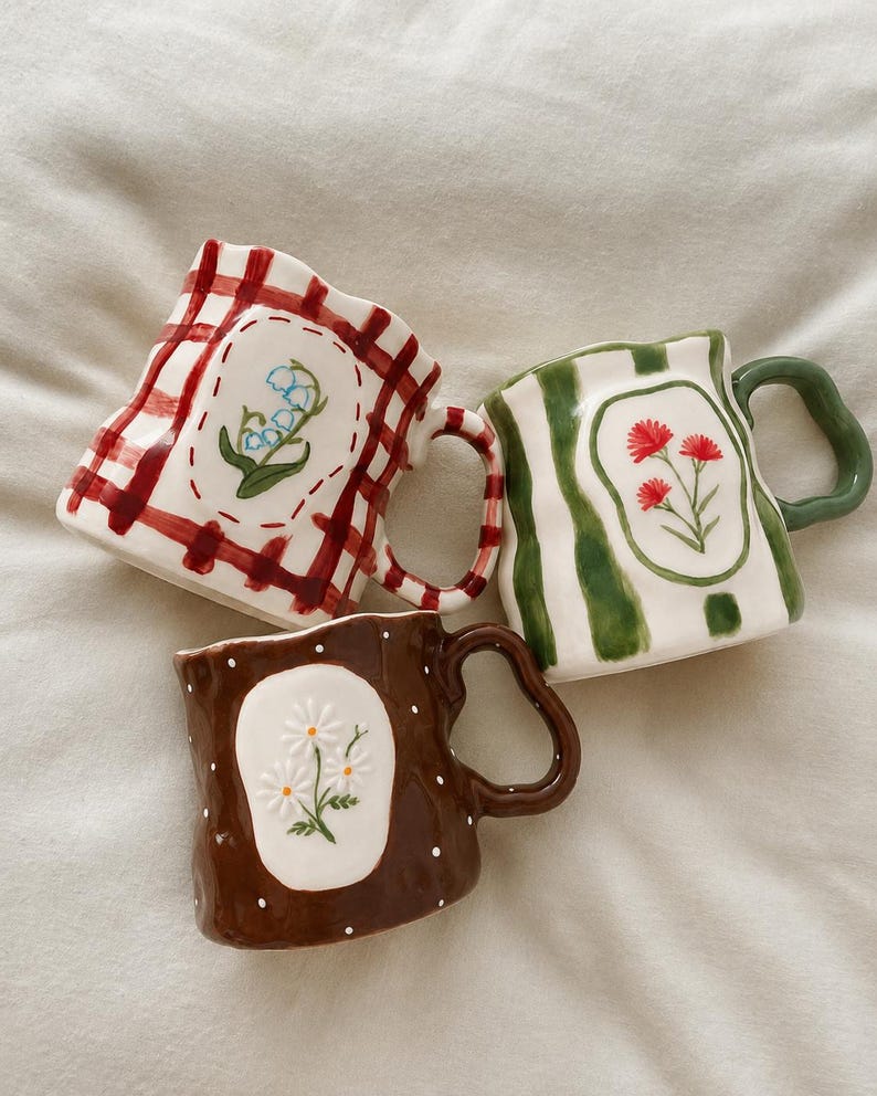May include: Three ceramic mugs with irregular, wavy rims and handles. One mug features a red gingham pattern with lily of the valley, one has green vertical stripes with red flowers, and one is brown with white polka dots and white daisies.