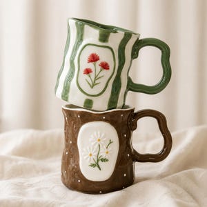 May include: Two ceramic mugs stacked vertically. The top mug is white with green vertical stripes and a red floral design. The bottom mug is brown with white polka dots and a white floral design. Both mugs feature wavy rims and handles.
