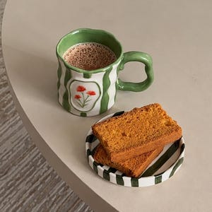 May include: A ceramic mug with a wavy rim and handle, featuring vertical green stripes and a central white panel with a small red flower illustration, filled with a brown beverage, next to a small round plate with green stripes holding two slices of toast.