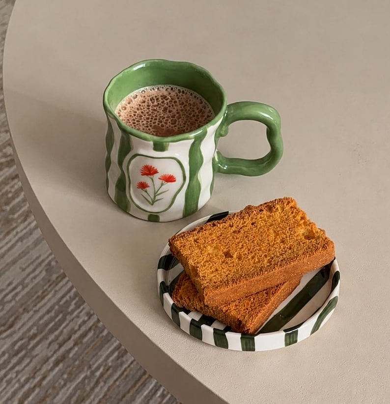 May include: A ceramic mug with a wavy rim and handle, featuring vertical green stripes and a central white panel with a small red flower illustration, filled with a brown beverage, next to a small round plate with green stripes holding two slices of toast.