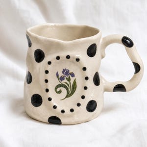 May include: A cream-colored ceramic mug with an irregular, hand-formed shape. The exterior features black polka dots and a central oval frame containing a painted purple iris flower with green leaves, surrounded by small black dots.