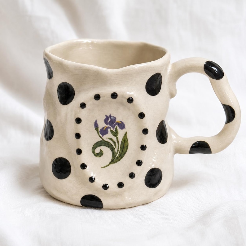 May include: A cream-colored ceramic mug with an irregular, hand-formed shape. The exterior features black polka dots and a central oval frame containing a painted purple iris flower with green leaves, surrounded by small black dots.