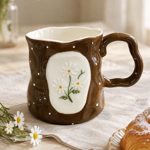 May include: A brown ceramic mug with a wavy, irregular rim and a matching wavy handle. The exterior features a glossy finish with small white polka dots and a central white oval panel displaying a raised, three-dimensional daisy flower design.