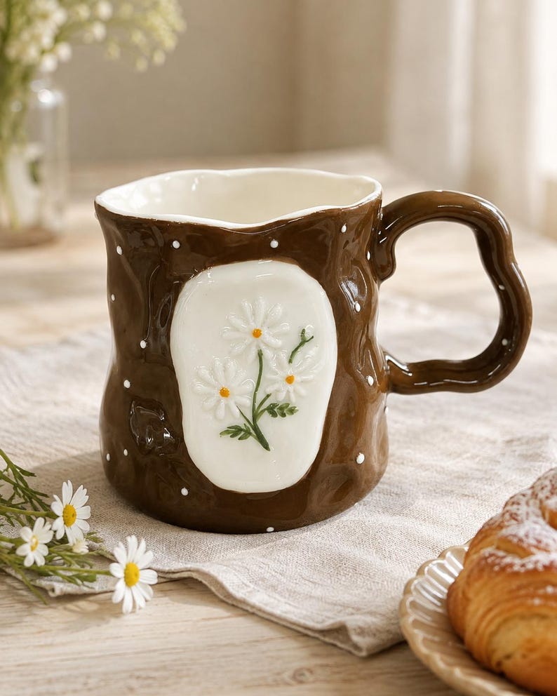 May include: A brown ceramic mug with a wavy, irregular rim and a matching wavy handle. The exterior features a glossy finish with small white polka dots and a central white oval panel displaying a raised, three-dimensional daisy flower design.