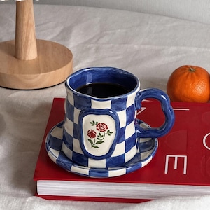 May include: A ceramic mug and matching saucer set featuring a blue and white checkered pattern. The mug has a wavy rim and a curved handle, with a central oval emblem displaying two red roses with green leaves. The mug is filled with dark liquid.