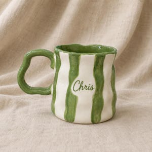 May include: A white ceramic mug with a wavy, irregular shape and a thick, green, hand-painted vertical striped pattern. The mug features a matching green handle and the name Chris written in a cursive green script on the front surface.
