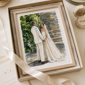 May include: A watercolour illustration of a couple standing together in wedding attire, framed in a light wood frame inside a gift box. The artwork depicts a person in a white suit and a person in a white gown with a veil, positioned before stone steps.