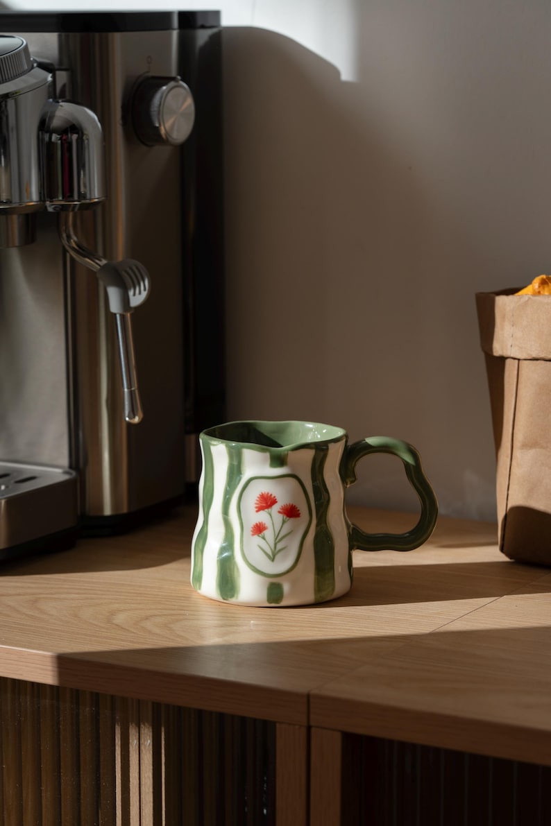 May include: A ceramic mug with a wavy, irregular rim and handle, featuring vertical green stripes on a white background. A central oval frame on the front displays a hand-painted illustration of three small red flowers with green stems.