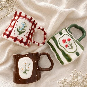 May include: Three ceramic mugs with irregular, wavy rims and handles. One mug features a red plaid pattern with a lily of the valley illustration, another has green stripes with red flowers, and the third is brown with white dots and daisies.