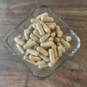 May include: A clear, square glass bowl filled with numerous beige capsules. The capsules are cylindrical with rounded ends, and the bowl sits on a wooden surface. The capsules appear to be a dietary supplement.