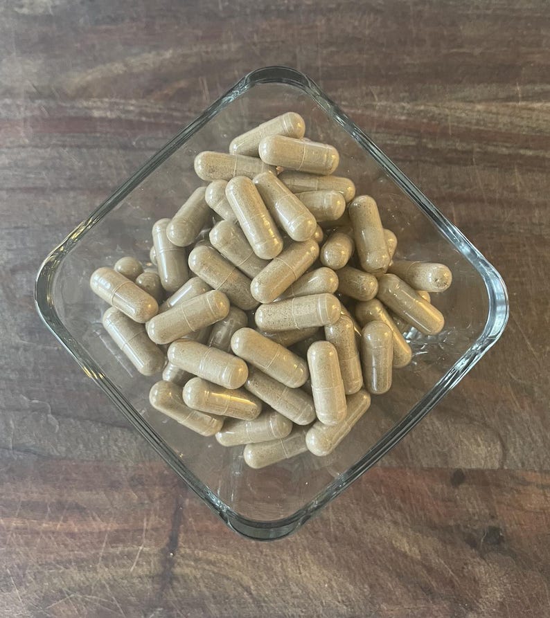 May include: A clear, square glass bowl filled with numerous beige capsules. The capsules are cylindrical with rounded ends, and the bowl sits on a wooden surface. The capsules appear to be a dietary supplement.