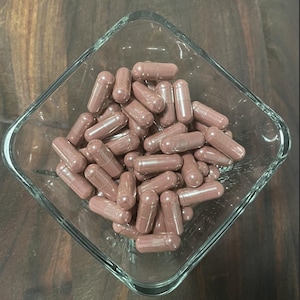 May include: A clear, square glass bowl filled with numerous maroon-colored capsules. The capsules are cylindrical with rounded ends, and they are densely packed within the bowl. The background is a dark, wooden surface.