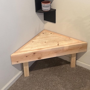 May include: A light-coloured, triangular wooden corner bench with two supporting legs. The bench is made of unfinished wood and is designed to fit snugly into a corner. The bench is placed on a carpeted floor.