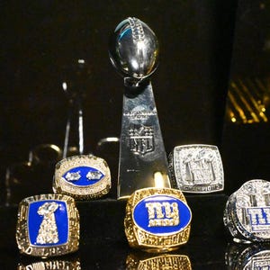 May include: A silver NFL Super Bowl trophy stands tall, surrounded by several gold and diamond-encrusted championship rings. The rings feature blue enamel and team logos, showcasing the team's victories.