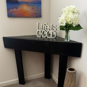 Puede incluir: Una mesa de esquina negra con tres patas, con un letrero blanco 'Life is Good', un jarrón con flores blancas y un jarrón decorativo. Una pintura de paisaje colorida cuelga de la pared encima de la mesa.