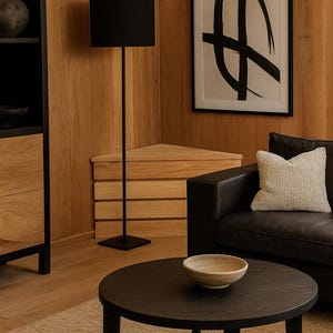 May include: A modern living room with wooden paneling and furniture. A black floor lamp with a rectangular shade stands near a wooden chest. A black round coffee table holds a bowl. A framed abstract art piece hangs on the wall.