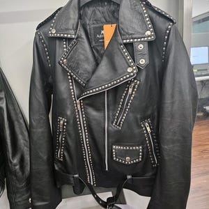 May include: Black leather jacket with silver stud accents. The jacket features a notched collar, asymmetrical zipper closure, multiple zippered pockets, and a belt at the waist. The jacket is hanging on a black hanger.