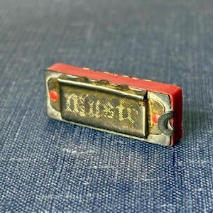 May include: A small, vintage harmonica with a red body and a silver-colored metal plate. The harmonica has the word "music" printed on it. The instrument is resting on a blue textured surface.