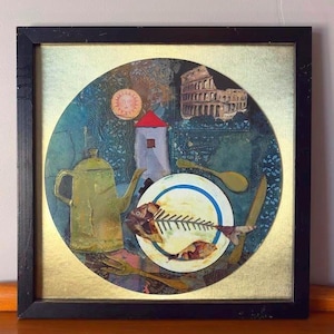 Framed mixed media collage artwork featuring a central white plate with a fish skeleton, surrounded by a golden teapot, wooden cutlery, and background motifs including a red-roofed tower, the Roman Colosseum, and a stylized sun. The frame is black with signs of wear, placed on a wooden surface against a light wall.