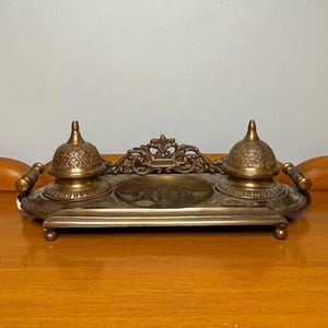 May include: An antique brass inkwell set. The set includes a rectangular tray with two ink pots, each topped with a domed lid. A decorative filigree piece sits between the ink pots. The tray has handles on each side.