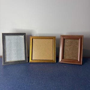 May include: Three rectangular picture frames arranged in a row. The left frame is black with a textured dotted pattern. The middle frame is square and gold-toned. The right frame is brown with a scalloped edge and a textured tan backing.