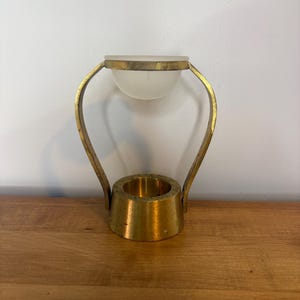 May include: A brass oil burner featuring a conical base with a central circular opening, connected by two curved brass arms to a suspended, frosted glass bowl at the top. The metal has a textured, aged finish with visible golden tones throughout.
