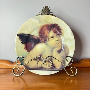 May include: A decorative plate featuring a cherub with red hair and a single wing, resting on a gold-colored metal stand. The plate has a cream-colored background with a cherub illustration. The stand has a pineapple-shaped top.