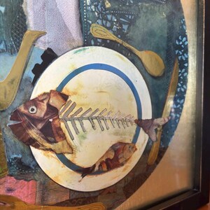 Zoomed-in view of the plate at the center of the artwork, featuring a detailed fish skeleton with collage textures. The tail end of the fish extends over the plate's edge. A small orange fish sits near the bottom of the plate, and gold cutlery surrounds it, adding depth and warmth to the composition.