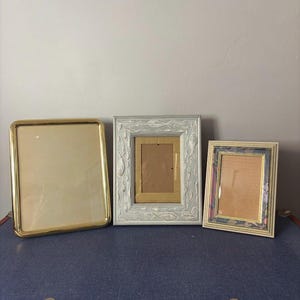 May include: Three decorative picture frames of varying sizes and styles. The frames include a rectangular gold-toned frame, a light blue ornate frame, and a smaller frame with a gold and patterned border. The frames are empty.